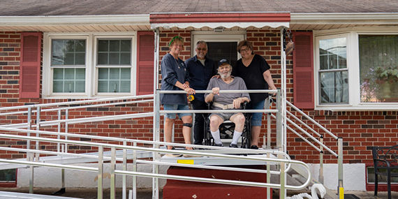 A senior disabled man sits in a wheelchair on his front porch surrounded by family