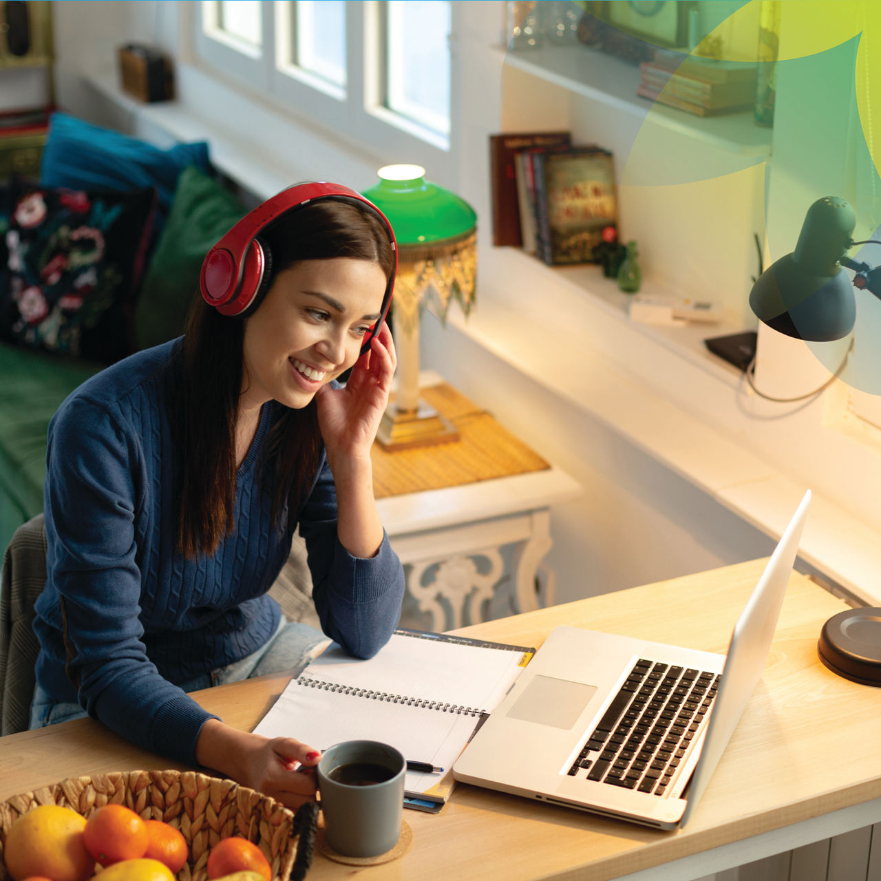 A young woman sits at her desk with a laptop and notepad, wearing headphones and holding a cup of coffee