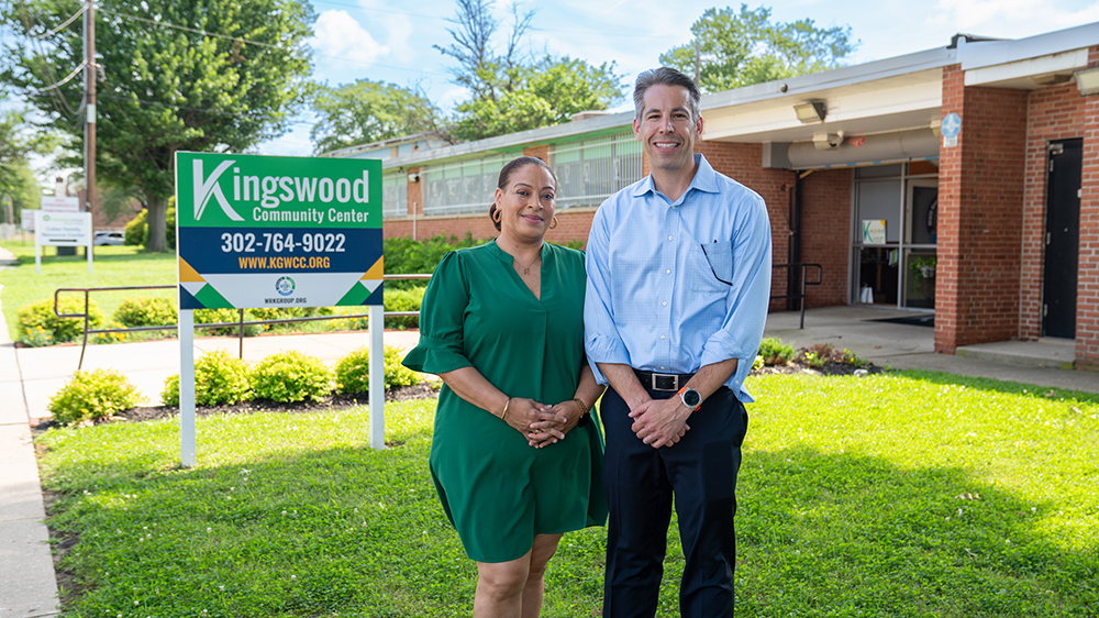 A man and a woman stand smiling in front of the Kingswood Community Center on a sunny day