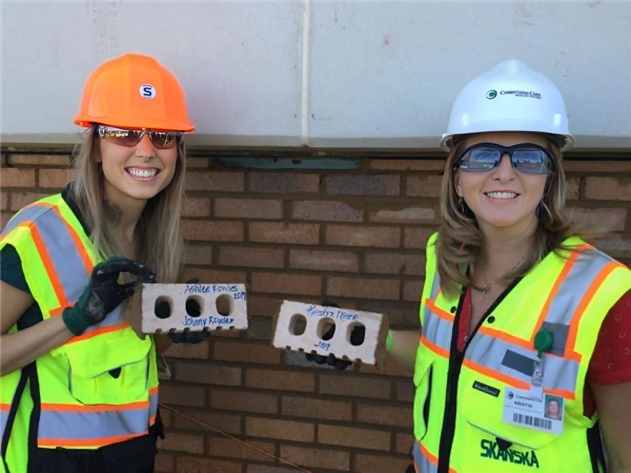 Two women wearing safety vests and construction helmets hold bricks