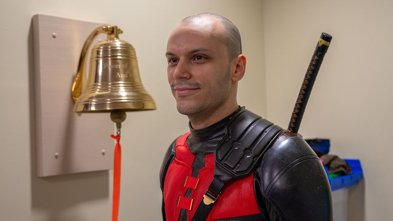 A patient dressed as Deadpool smiles beside the cancer center's ceremonial bell, marking the completion of treatment.