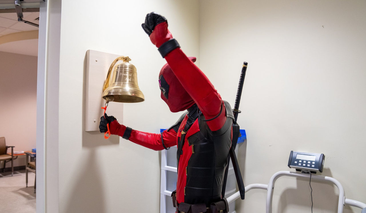 A patient in a Deadpool costume raises a fist in triumph while ringing the cancer center's victory bell.