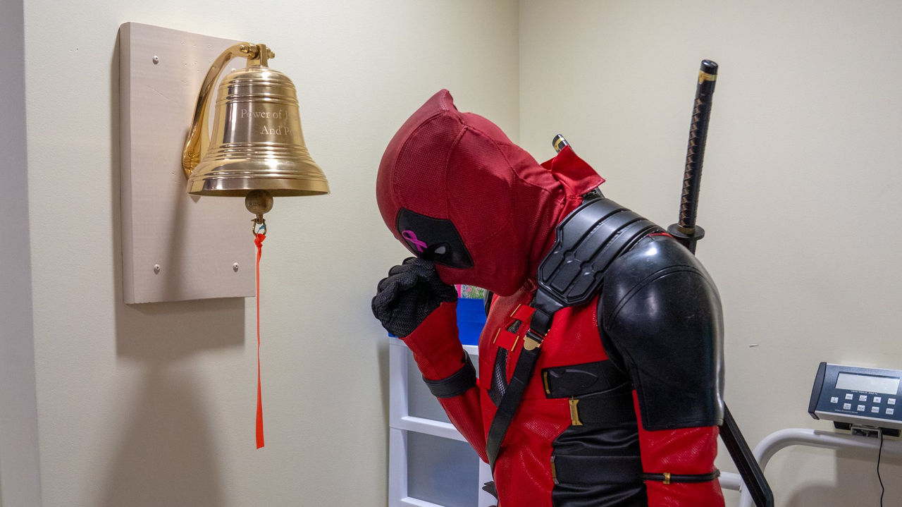 A patient in a Deadpool costume pauses in a reflective moment before the cancer center's victory bell.