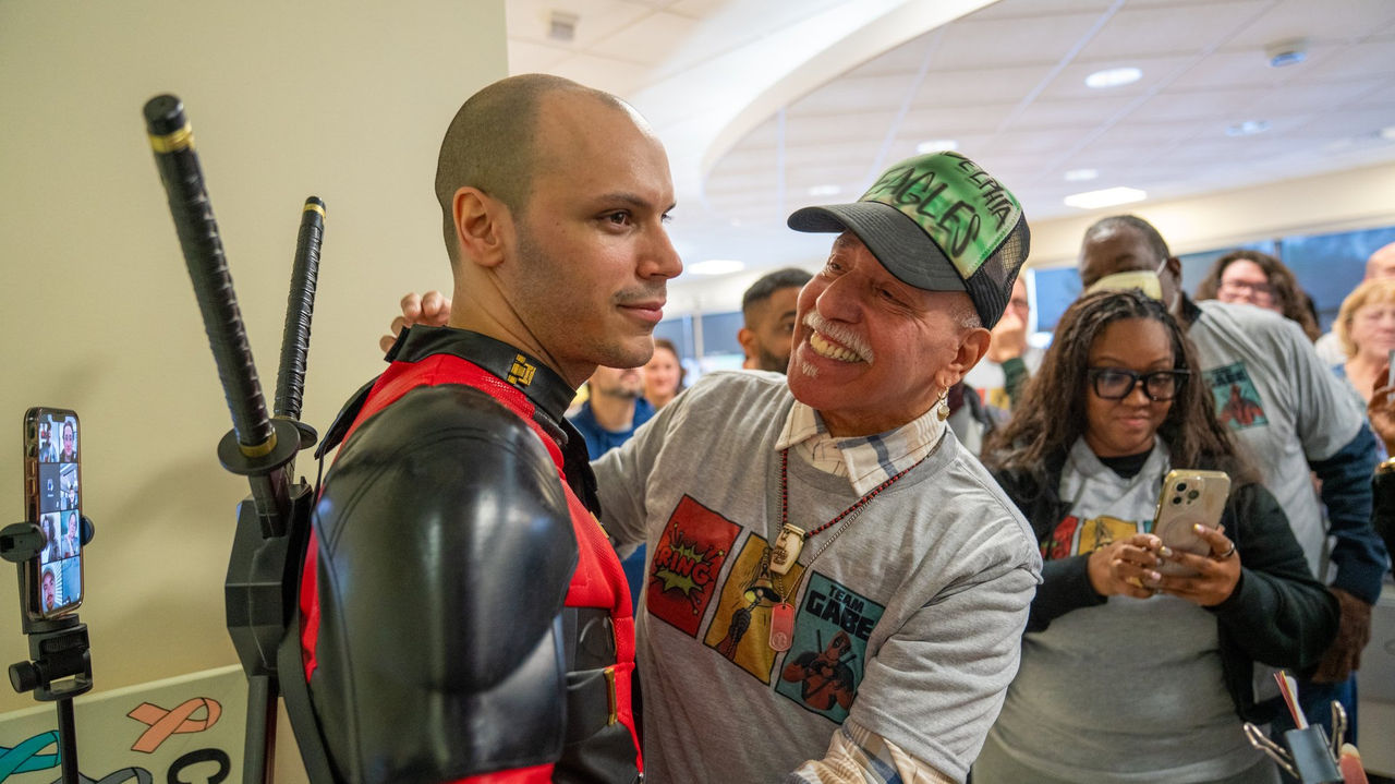 A patient in a Deadpool costume shares a warm embrace with a fellow cancer center visitor during the bell-ringing celebration.