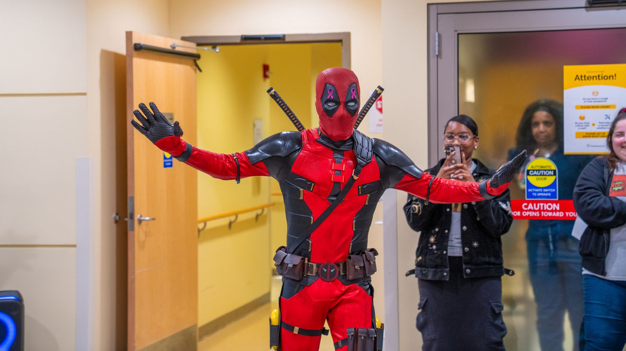 A patient in a full Deadpool costume strikes a dramatic pose in the hallway as staff look on with smiles at the cancer center.