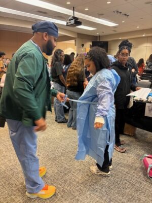 A student is helped into a surgical gown by a clinical staff member during a hands-on career fair activity.