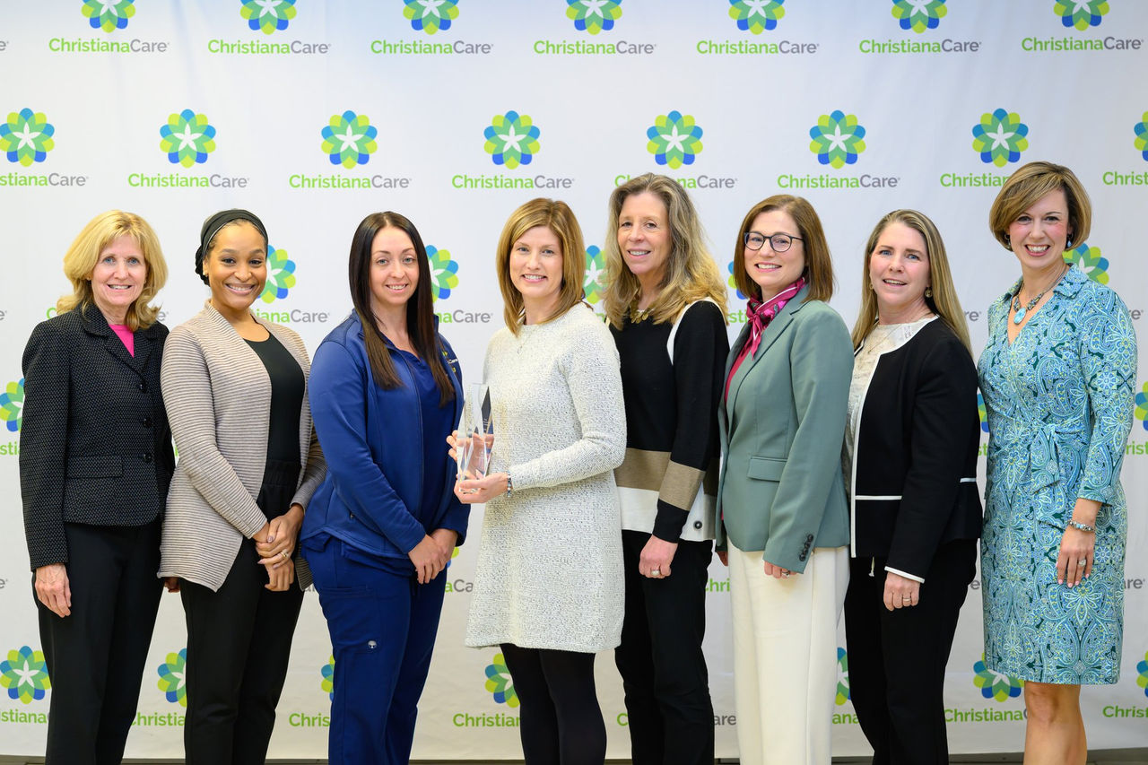 A group of eight colleagues poses with a crystal trophy in front of a ChristianaCare branded backdrop at the Way Awards ceremony.
