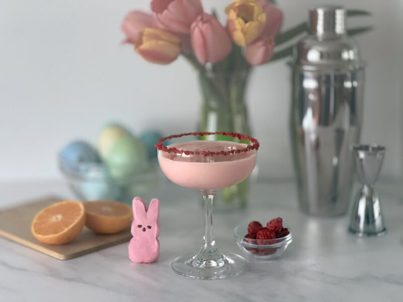 A creamy pink raspberry cocktail in a red sugar-rimmed coupe glass, nestled beside a pink Peeps bunny, fresh raspberries, and halved oranges — styled with pastel Easter eggs, pink tulips, and a cocktail shaker for a delightful spring celebration drink.