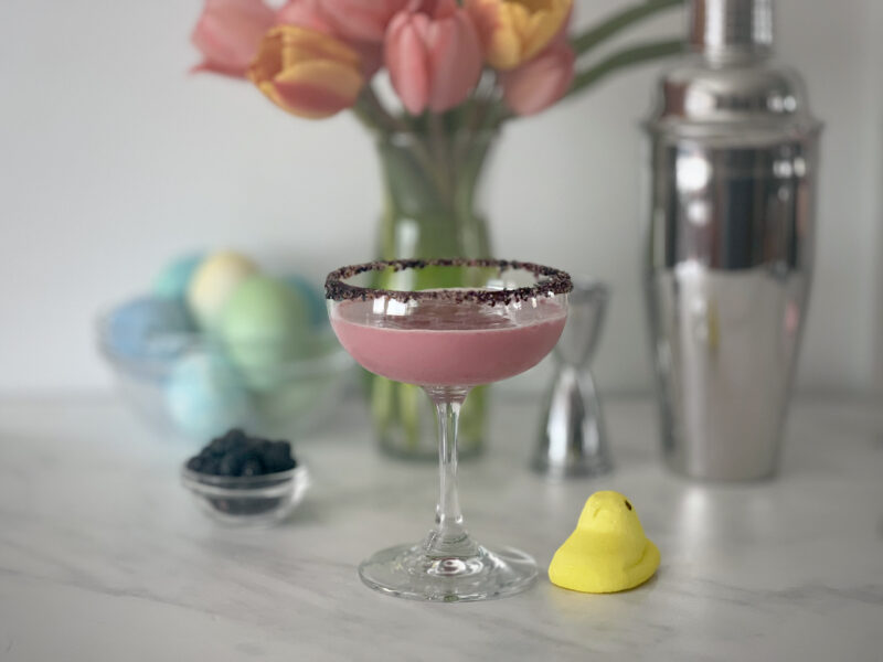 A soft lavender blueberry cocktail in a dark sugar-rimmed coupe glass, accompanied by a yellow Peeps chick and a small bowl of blueberries — styled with pastel Easter eggs, pink tulips, and a cocktail shaker for a charming spring drink setup.