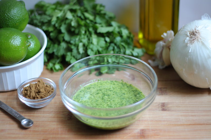 A clear glass bowl filled with a blended green herb marinade sits centered on a wooden cutting board in image_87969a.jpg. The surrounding ingredients include a bunch of fresh cilantro, a bowl of whole limes, a large white onion, garlic cloves, a bottle of olive oil, and a small dish of ground cumin with a measuring spoon.