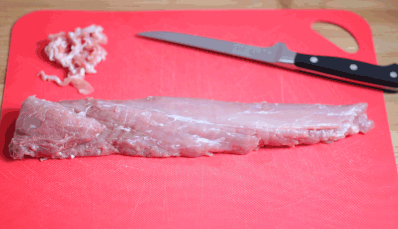 Two long strips of raw pork tenderloin lie horizontally across a bright red cutting board in image_8799be.png. A pile of trimmed fat sits in the upper left corner, and a sharp utility knife rests in the top right corner.