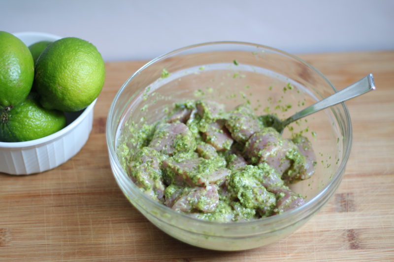A clear glass bowl filled with cubed meat marinating in a vibrant green herb mixture sits on a wooden cutting board next to a small white bowl of whole limes