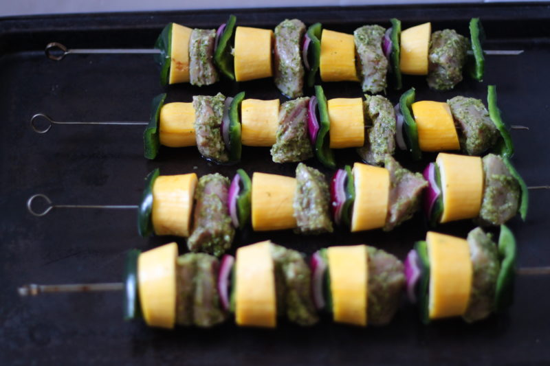 Uncooked skewers layered with marinated pork, yellow squash, green peppers, and red onion are arranged on a baking tray, ready for grilling or roasting.