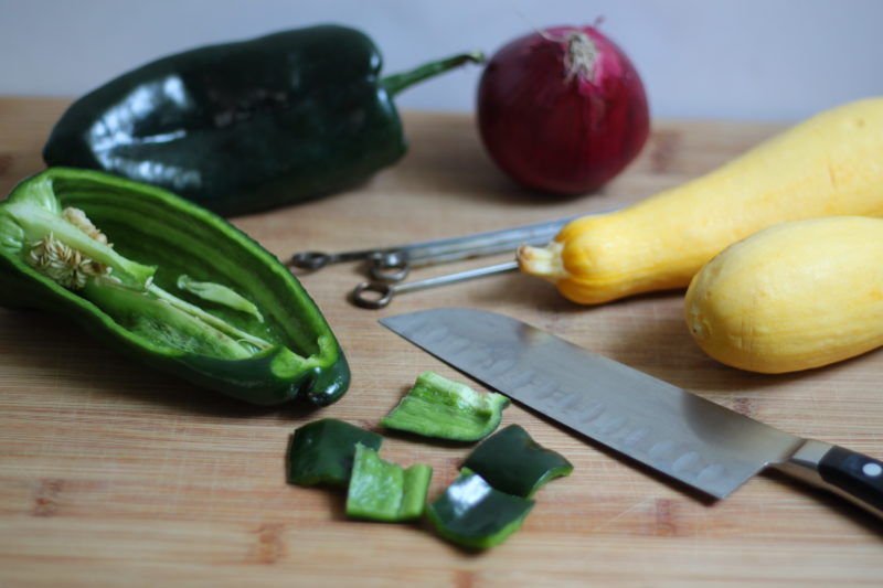 A light wooden cutting board displays fresh ingredients being prepped for cooking. It features a sliced poblano pepper with scattered chopped pieces, a Santoku-style chef's knife, a whole red onion, two yellow summer squashes, and a few metal skewers ready for grilling.