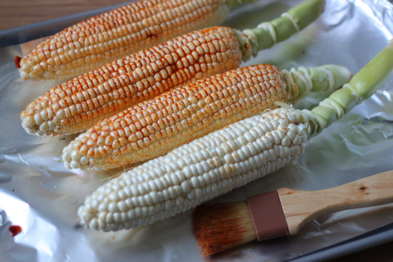 Fresh ears of sweet corn laid out on a foil-lined baking sheet, getting brushed with a rich, smoky chili seasoning. A rustic and delicious look at the prep work behind a flavorful, oven-roasted treat.