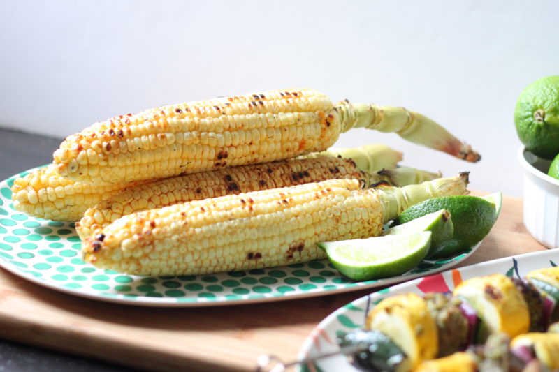 Charred ears of corn stacked on a teal polka-dot plate with fresh lime wedges — a simple, smoky summer side dish served alongside herb-marinated skewers for a vibrant backyard cookout spread.