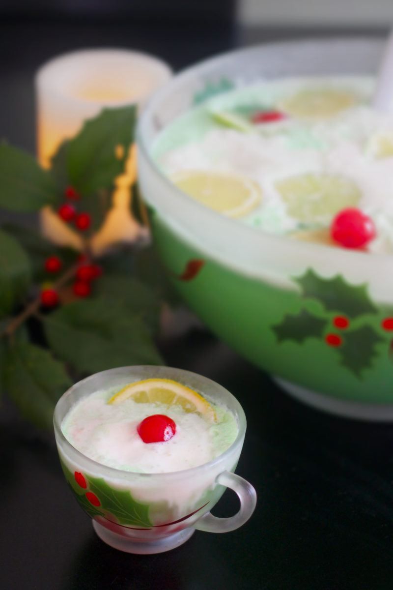 A frothy green holiday punch served in a vintage holly-print cup, garnished with a lemon slice and a maraschino cherry — with a matching festive punch bowl in the background and a sprig of red-berry holly adding a classic Christmas touch.
