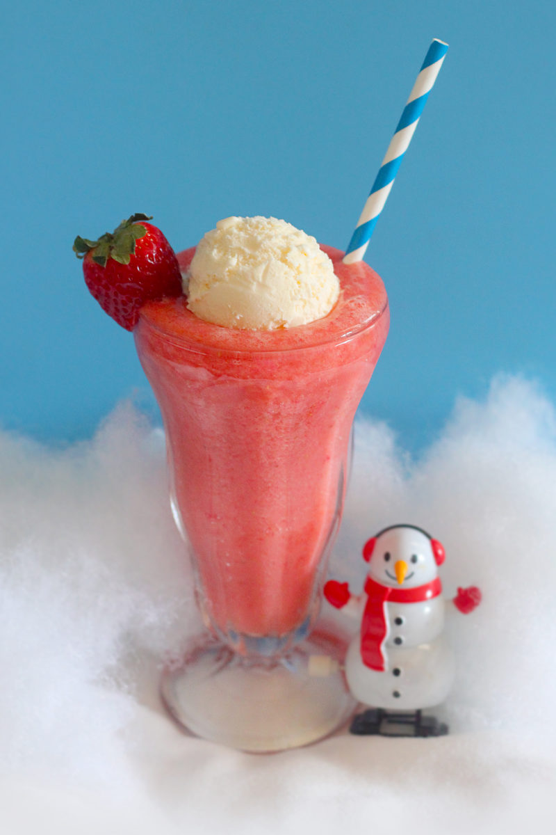 A vibrant pink strawberry slush topped with a scoop of vanilla ice cream and a fresh strawberry, served with a blue striped straw — nestled in fluffy artificial snow beside a cheerful snowman figurine against a bright blue winter backdrop.