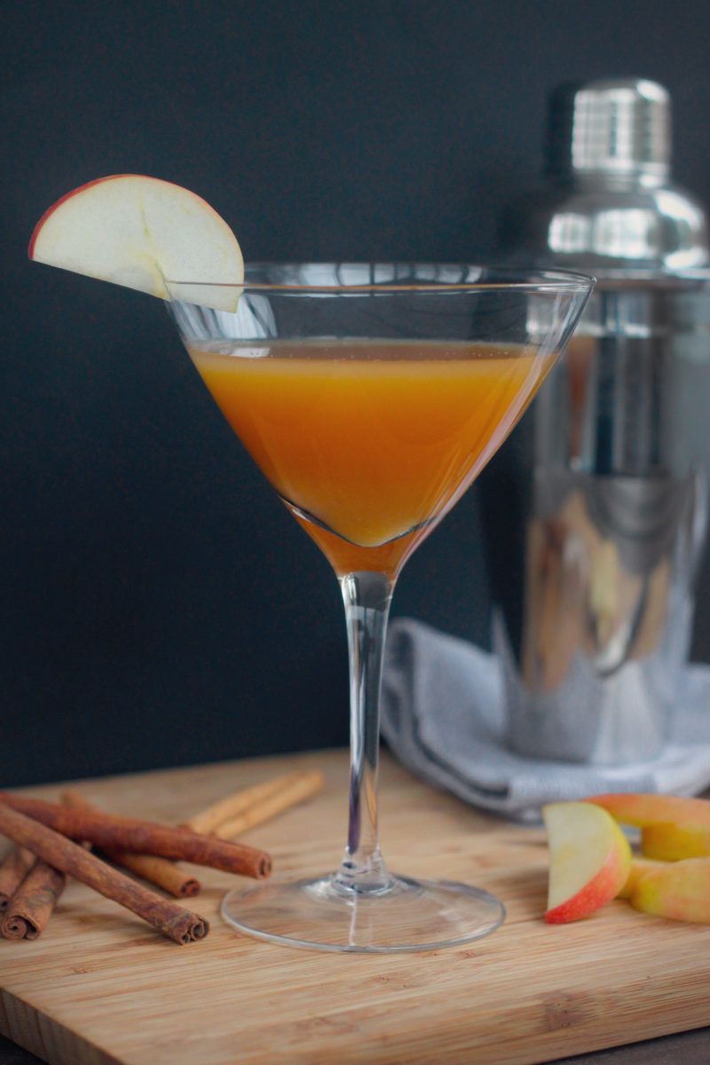A golden-amber apple martini garnished with a thin apple slice, served in a classic martini glass on a wooden board surrounded by cinnamon sticks and fresh apple wedges — with a cocktail shaker softly blurred in the background.