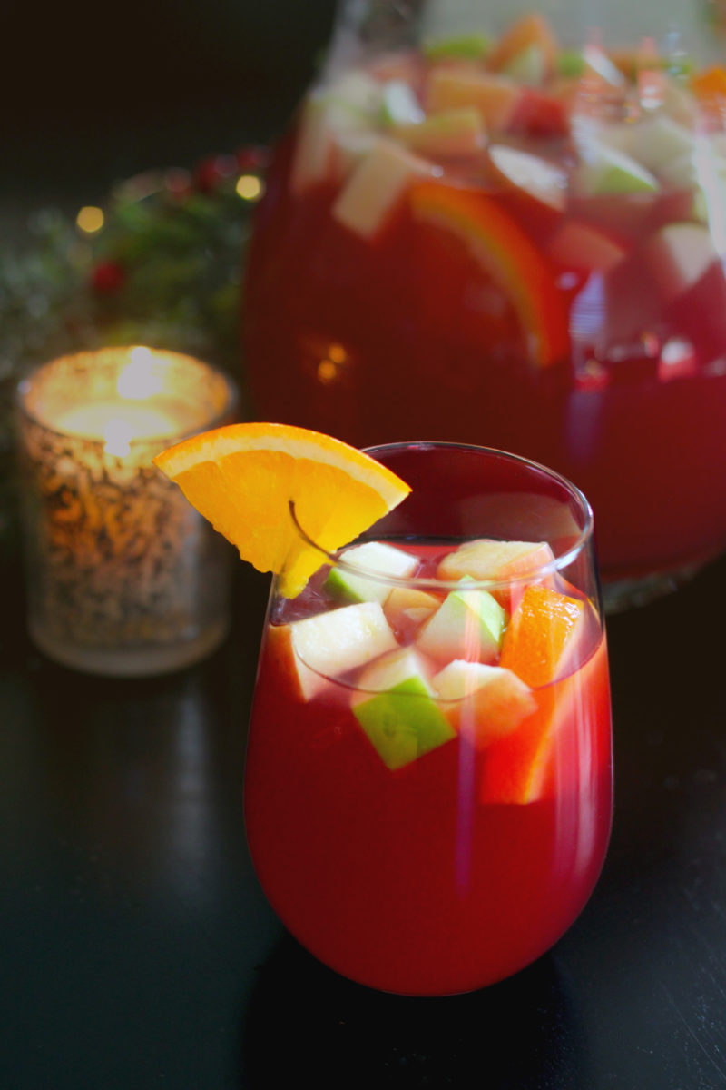 A deep ruby sangria filled with chopped apple, orange, and pear, garnished with an orange wedge — set against a moody candlelit backdrop with a pitcher in the background, evoking a warm festive atmosphere.