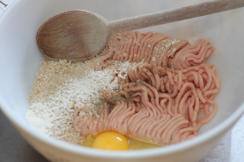 A large white ceramic bowl contains the unmixed ingredients for a meatball or meatloaf recipe in image_790b34.jpg. Inside, a mound of ground pink meat sits alongside a pile of breadcrumbs, a dusting of flour or seasoning, and a raw whole egg yolk, with a wooden spoon resting along the top rim ready to stir.