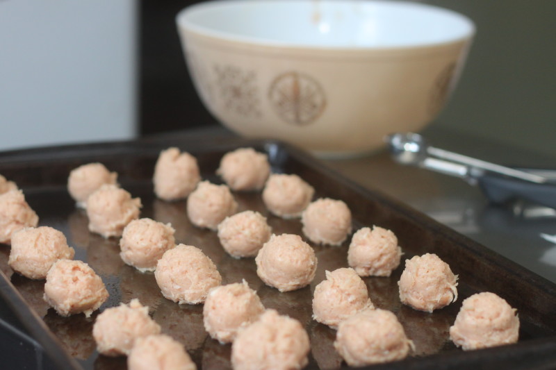 A dark baking sheet filled with neatly arranged rows of small, raw pink meatballs dominates the foreground in image_790ea1.jpg. In the blurred background, a patterned ceramic mixing bowl and a metal cookie scoop rest on the counter, showing the process of shaping the mixture.