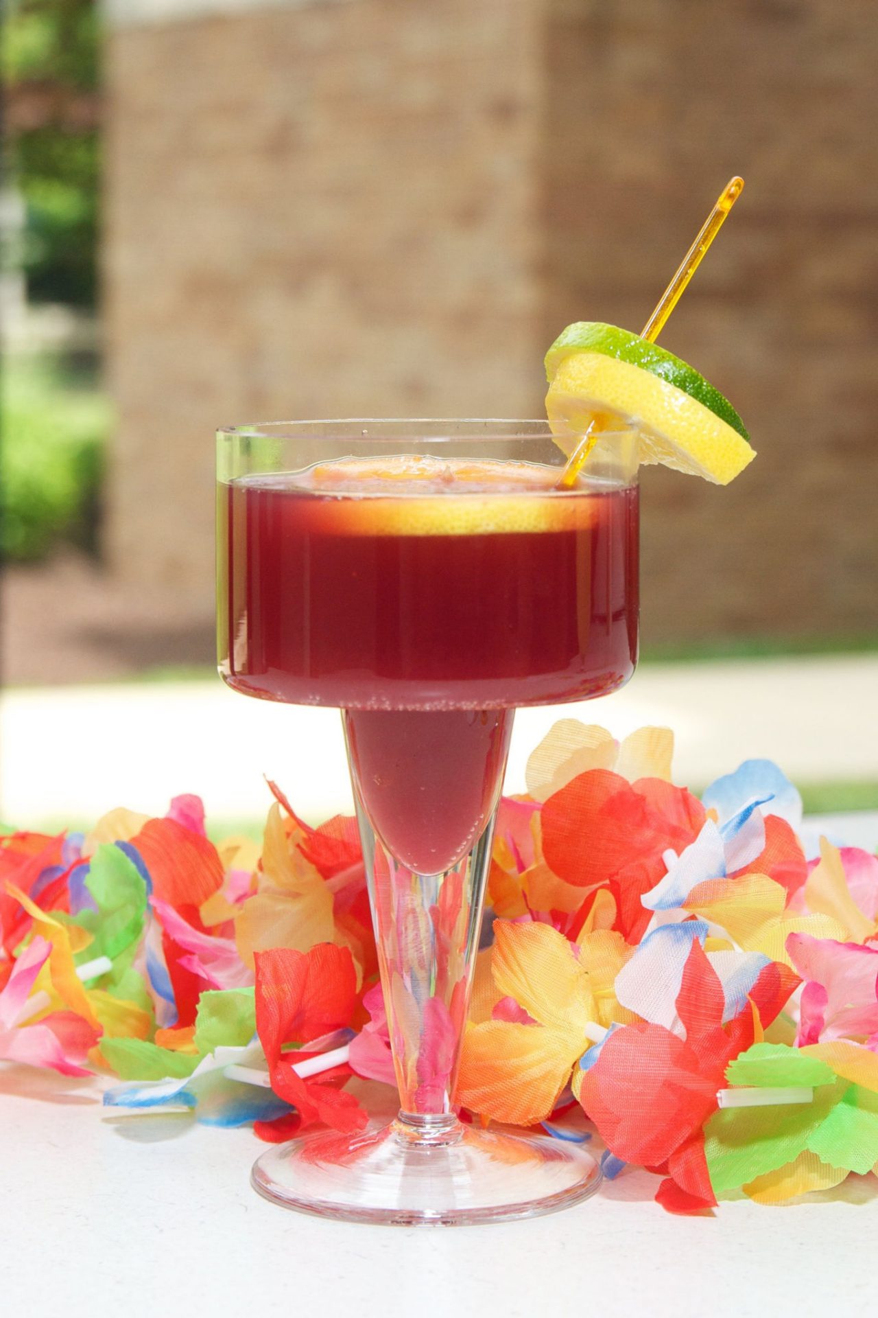 A wide-rimmed coupe glass filled with a deep red fruit punch or cocktail is shown in image_796458.jpg. The drink contains a floating orange slice and is garnished on the rim with stacked lemon and lime wheels pierced by a yellow stirrer, all set against a vibrant, multi-colored flower lei on the table.