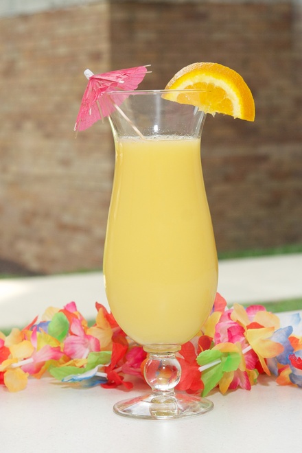 A tall hurricane glass filled with a bright yellow citrus drink is featured in image_7964d3.jpg. The beverage is garnished with an orange slice on the rim and a small pink cocktail umbrella, while a colorful Hawaiian-style flower lei lies on the surface behind the base of the glass.